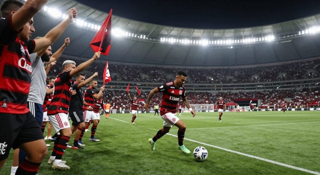 A dynamic football scene at a bustling stadium, capturing the essence of a match featuring Flamengo's iconic red and black colors. In the foreground, an enthusiastic group of Flamengo fans, dressed in team jerseys, wave flags and create an electric atmosphere. In the middle ground, a sharp action shot of Kaio Jorge, the attacking player, skillfully dribbling the ball, focused and determined, with the stadium crowd cheering behind him. The background showcases the vibrant stadium lights illuminating the field, casting long shadows and enhancing the sense of movement. The mood is spirited and hopeful, reflecting the excitement of the crowd and the significance of the moment. The image should be captured from a slightly low-angle perspective to emphasize the players and fans, with soft yet dramatic lighting to create depth.