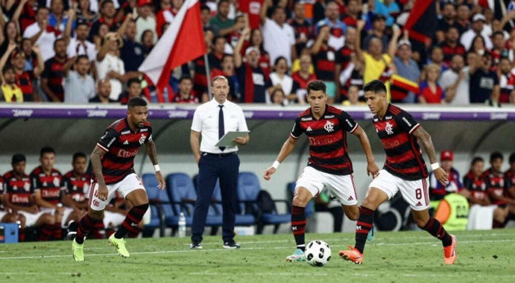 A tactical football scene capturing the essence of the Flamengo matches from 2019, focusing on their dynamic playing style. In the foreground, two players in professional football kits are engaged in a thrilling play, one dribbling the ball past the other, showcasing agility and teamwork. In the middle ground, a coach in business attire analyzes the game, clipboard in hand, reflecting strategic thinking. The background features an energetic crowd in a stadium, filled with vibrant team colors and flags waving. The lighting is bright and dynamic, highlighting the players and creating a sense of action. The atmosphere is charged with excitement, embodying passion and competitive spirit typical of high-stakes football matches.