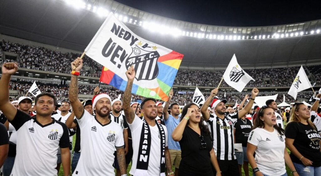 A vibrant and dynamic scene capturing the passionate reception of the Santa fans as Neymar returns to Santos in 2026. In the foreground, a diverse group of enthusiastic fans, wearing the team's colors—white and black—cheer and wave flags, radiating excitement. Their attire consists of casual jerseys and scarves. The middle ground depicts a large, colorful banner with Neymar's name and number, symbolizing their support and anticipation. In the background, the iconic Estádio Urbano Caldeira is packed with more supporters, creating a sea of energy and unity. Bright stadium lights illuminate the scene, casting a warm glow over the celebration, enhancing the atmosphere of joy and hope. The perspective is slightly low-angle, emphasizing the fans' enthusiasm and the grand moment of Neymar’s return.